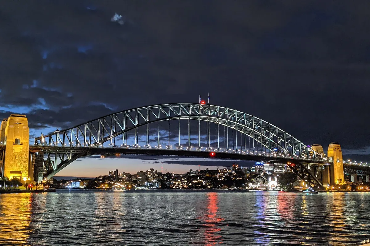 El Sydney Harbour Bridge de noche, con su arco y sus pilonos de arenisca iluminados reflejados en el puerto, y las luces de la orilla norte al fondo.