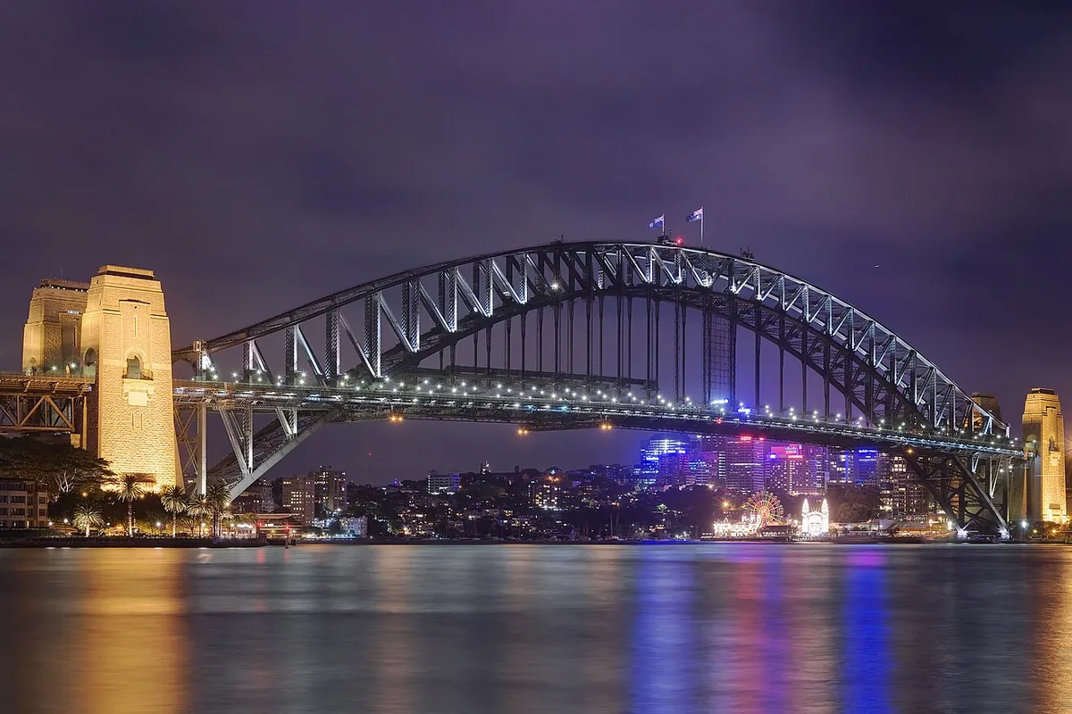 El Sydney Harbour Bridge de noche visto desde Circular Quay, con su arco perfilado por luces contra un cielo morado y un pilono de arenisca a la izquierda.