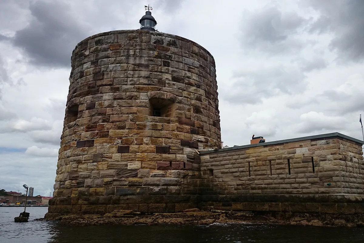 La torre redonda de arenisca Martello de Fort Denison vista desde el agua, con un farol en lo alto y una baja muralla de piedra que se extiende hacia la derecha.
