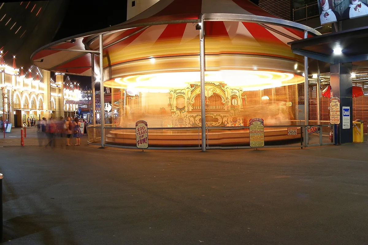 Un carrusel con toldo a rayas rojas y blancas que brilla de noche en Luna Park Sydney, con las luces engarzadas de un edificio a la izquierda.