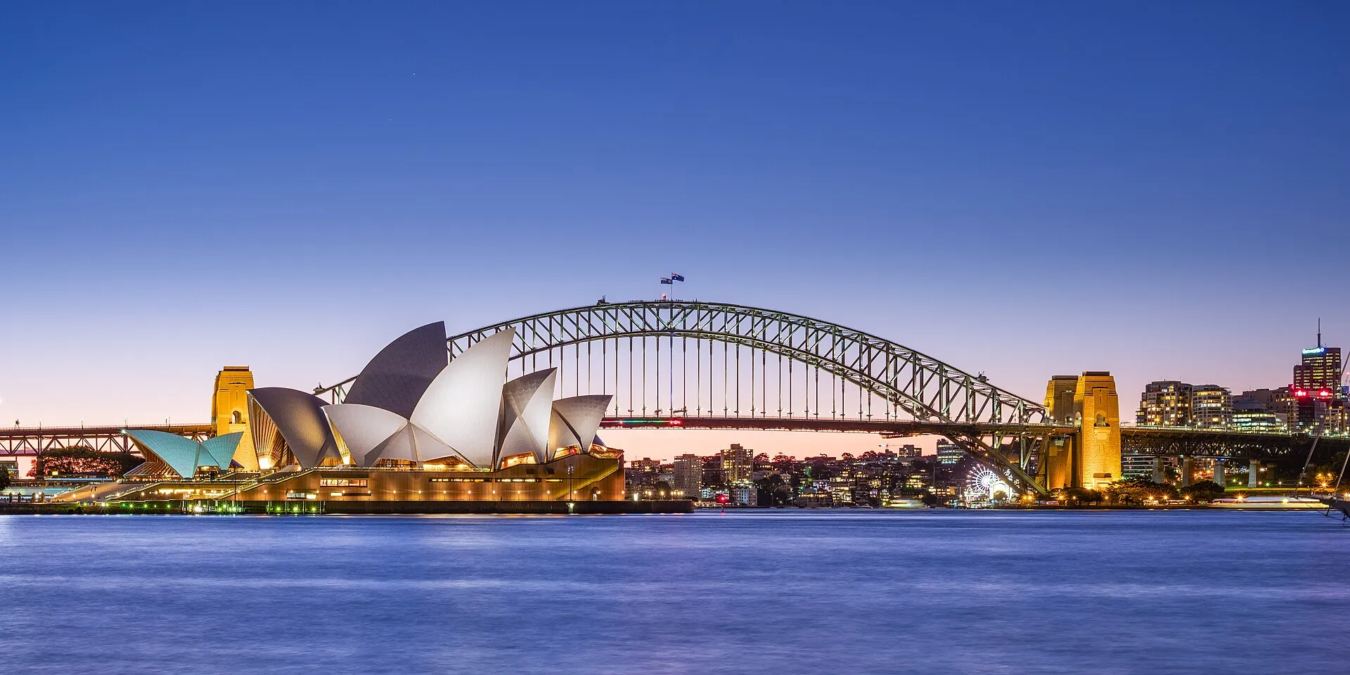 La Sydney Opera House y el Sydney Harbour Bridge al atardecer, con las velas iluminadas de la Ópera y el arco de acero del puente sobre las tranquilas aguas azules.
