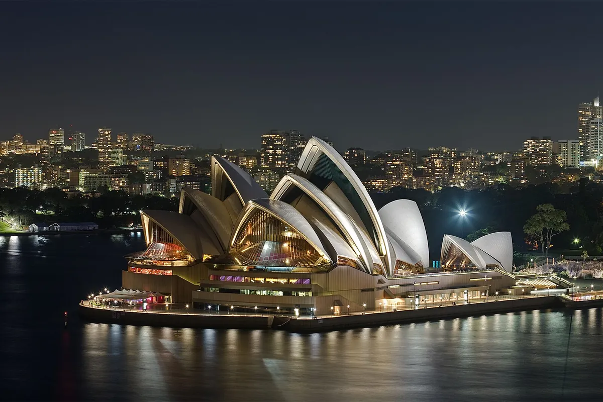 La Sydney Opera House iluminada de noche, con sus velas blancas y sus extremos de cristal resplandecientes sobre el puerto oscuro, y las luces de la ciudad al fondo.