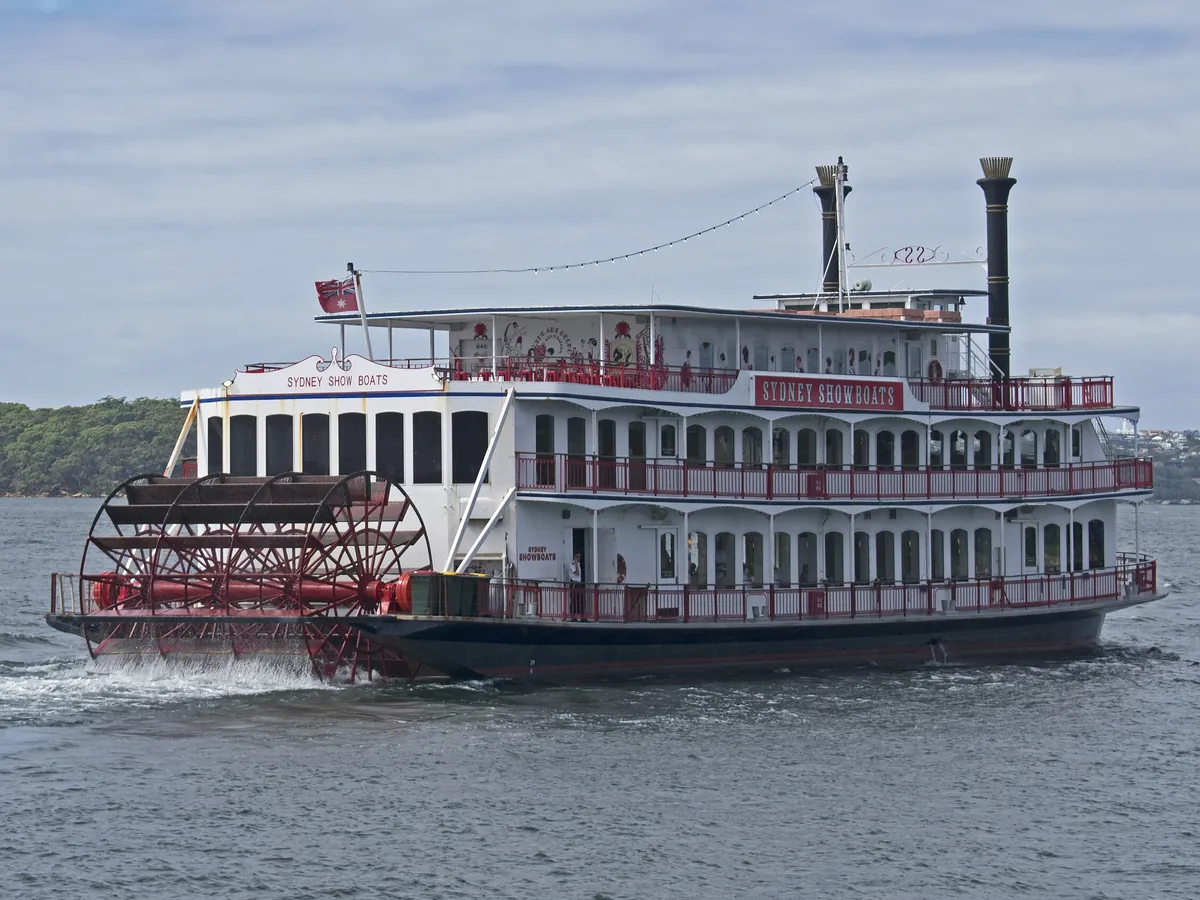 El showboat de rueda de paletas roja y blanca Sydney Showboat II en el puerto de Sídney a la luz del día, con su rueda de paletas roja en la popa, pisos de cubiertas blancas con barandillas rojas y dos chimeneas negras.
