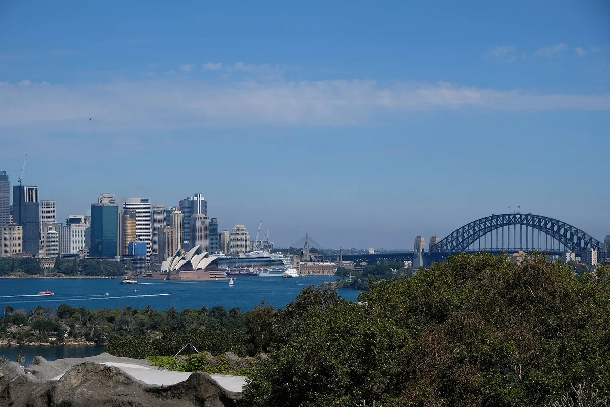 Una vista del puerto de Sídney desde la orilla del zoológico de Taronga hacia las torres de la ciudad, la Ópera y el Harbour Bridge, con vegetación verde en primer plano.