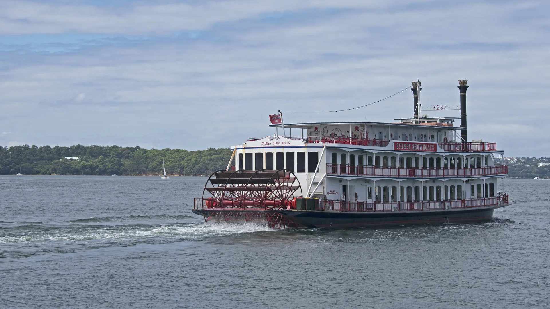 El barco de espectáculos de ruedas de paletas rojo y blanco Sydney Showboat II navegando por el puerto de Sídney, con su rueda de paletas roja en la popa y dos chimeneas negras sobre la cubierta superior.